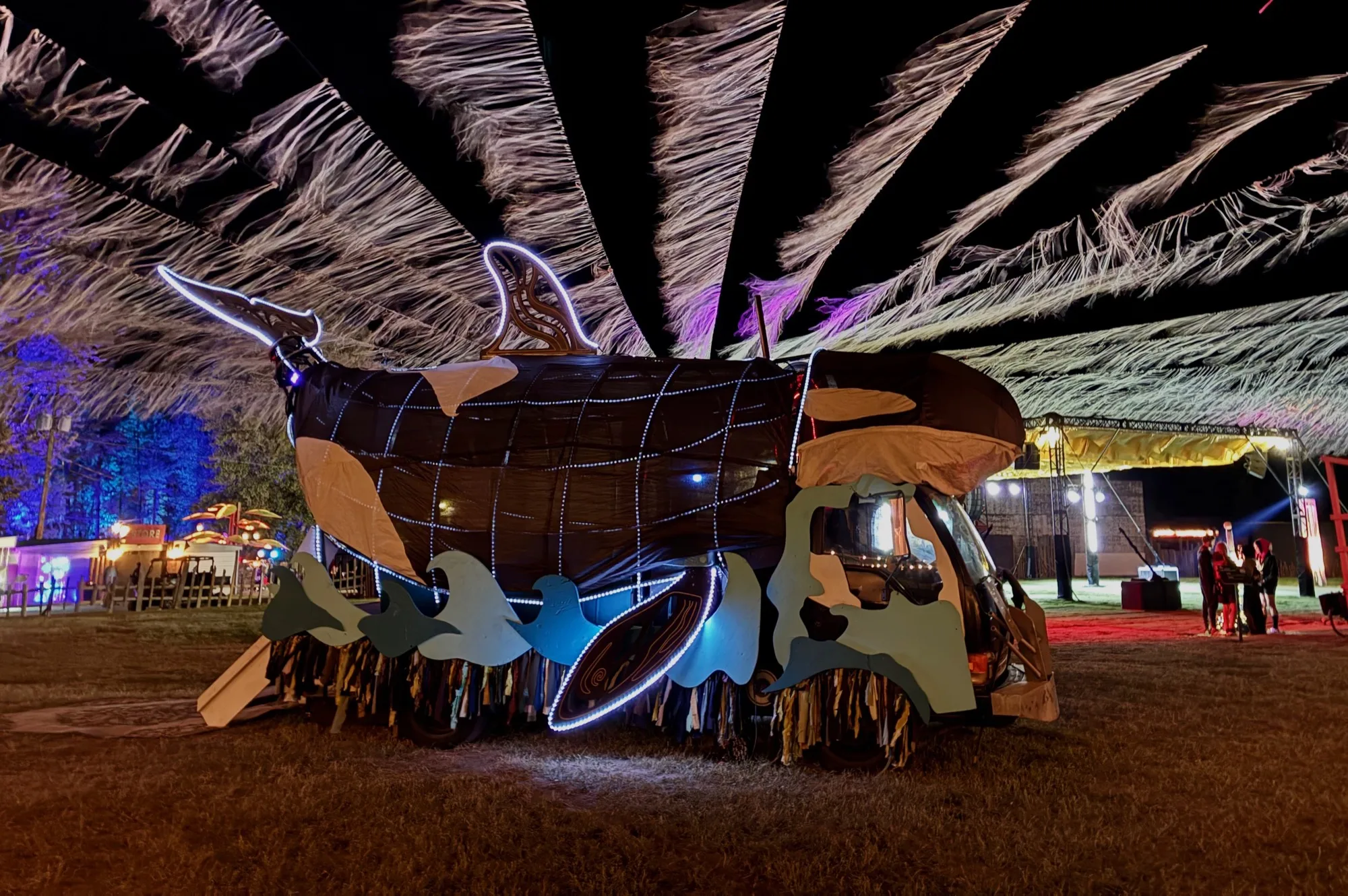The Orcar parked beneath a fringed festival canopy at night, white LED rope tracing the orca outline
