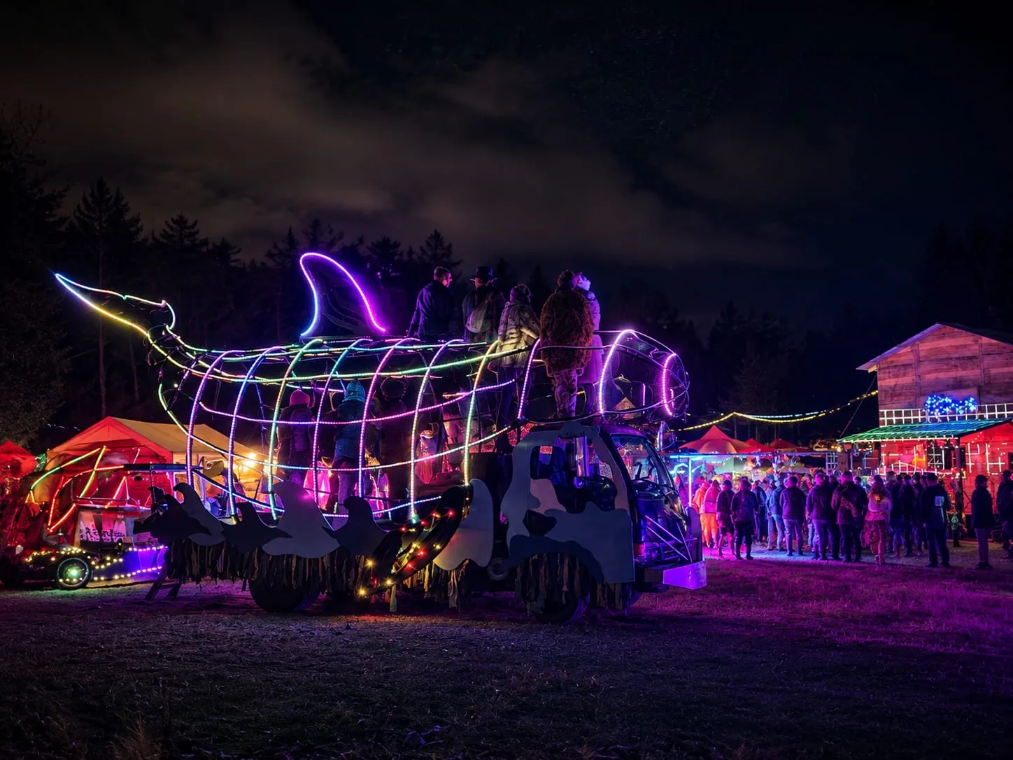 The Orcar at night, its orca rib-cage traced in rainbow LED rope, riders standing aboard as a festival crowd gathers behind