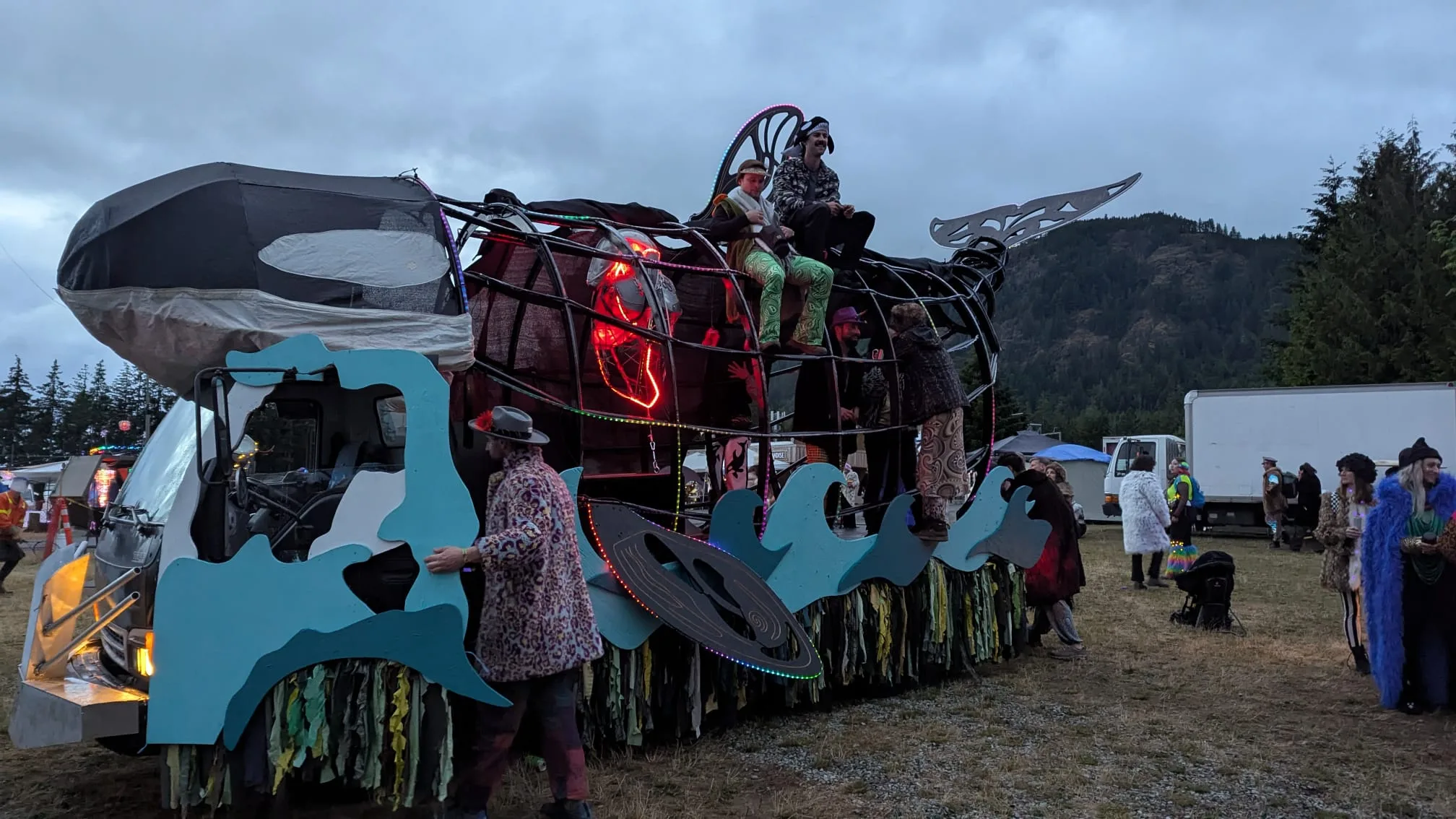 The Orcar at dusk with riders aboard, a forested mountain ridge and festival grounds behind