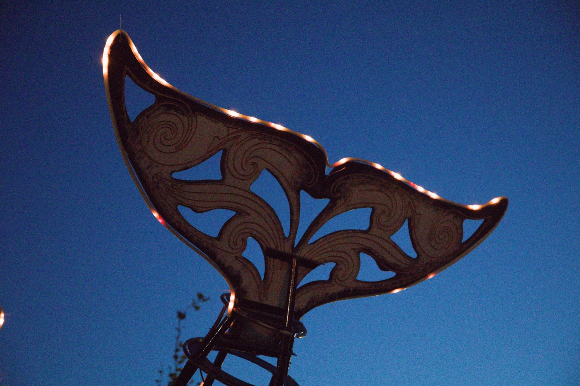 Detail of the Orcar tail fluke, laser-cut steel with carved wave scrollwork, LED-edged against a deep blue dusk sky