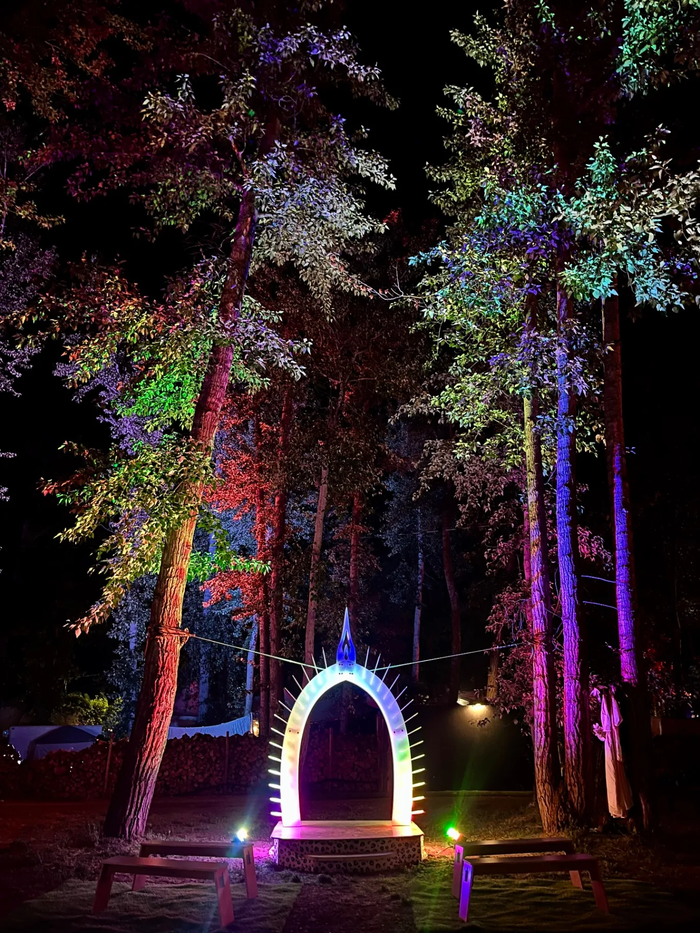 The Arch at night — the illuminated ceremony arch glowing on its raised platform, benches set before it and the surrounding forest washed in rainbow light