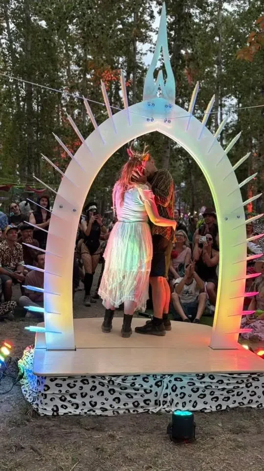 A couple kisses under the Arch at Bass Coast, the arch glowing in rainbow light as the crowd watches