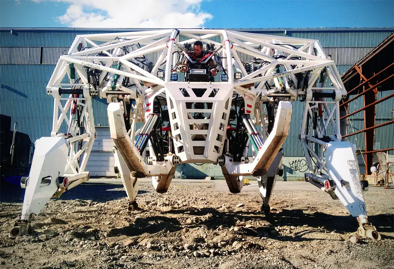 Prosthesis, a white four-legged exoskeleton mech, standing with its legs extended on open ground under a blue sky