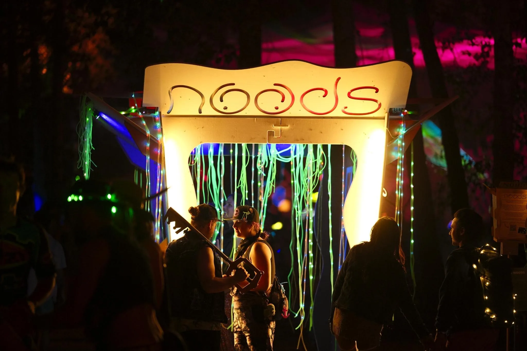 Festival-goers crowd around the lit Noodle Box at night, watching someone framed behind the noodle curtain