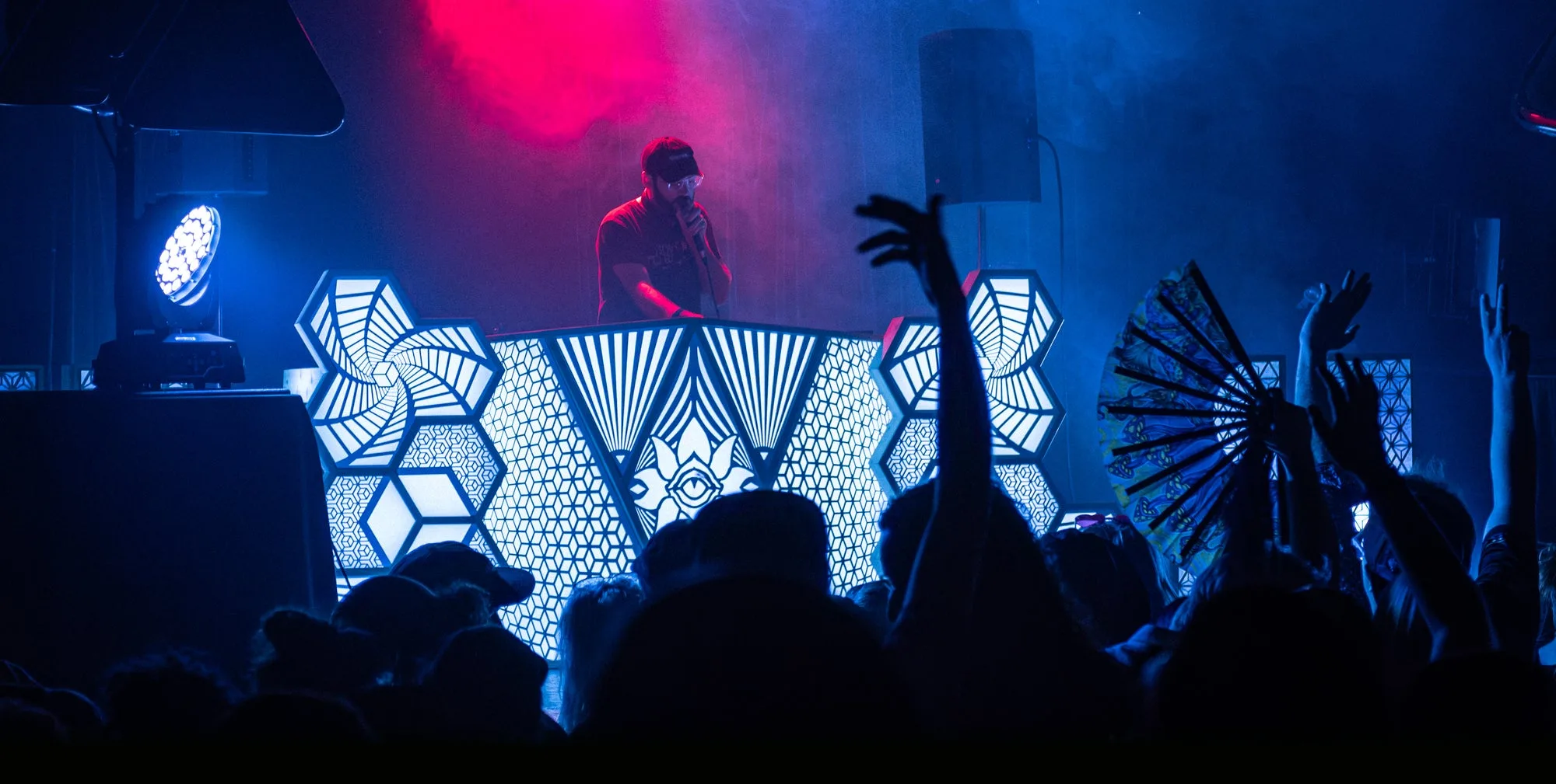 A DJ performing behind The Observer at a club show, its white-lit panels glowing over a crowd with raised hands