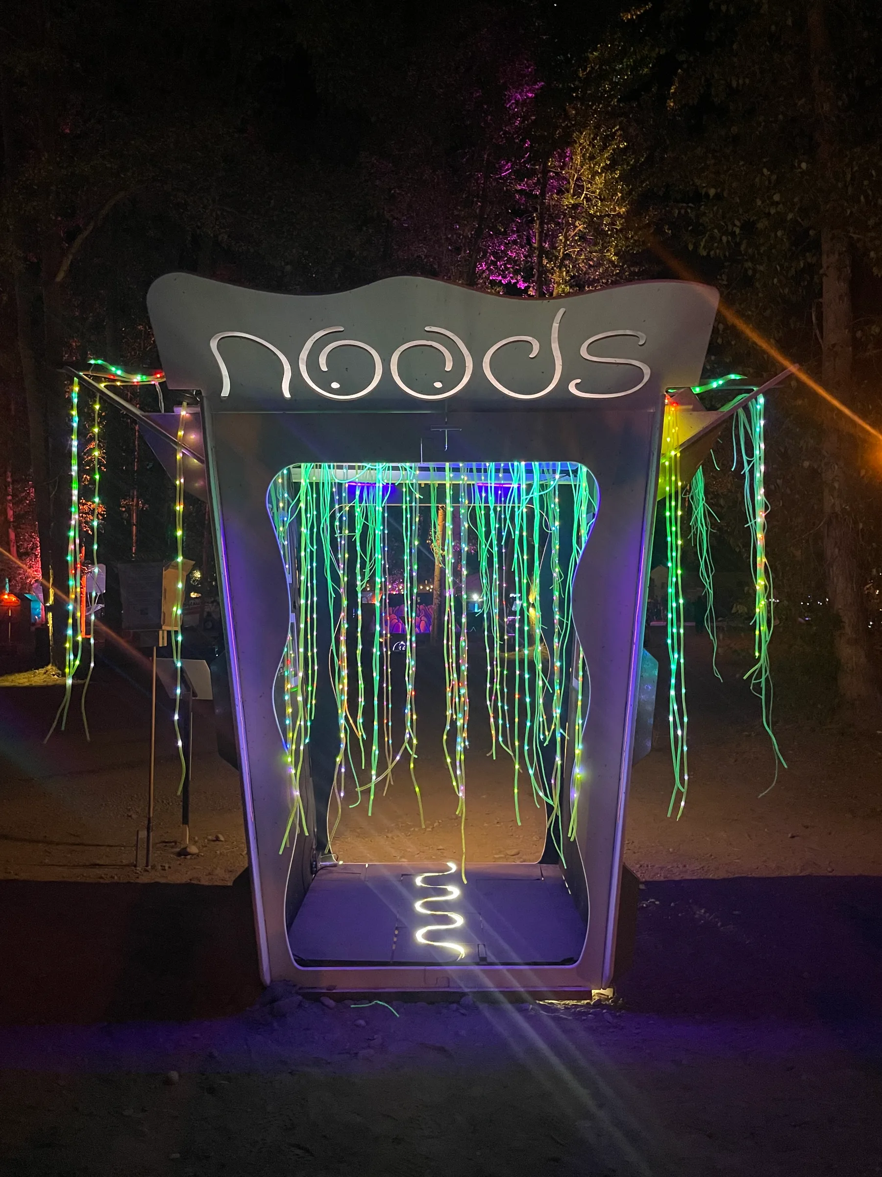 The Noodle Box in a forest clearing at night, rainbow LED noodles hanging from the open lid