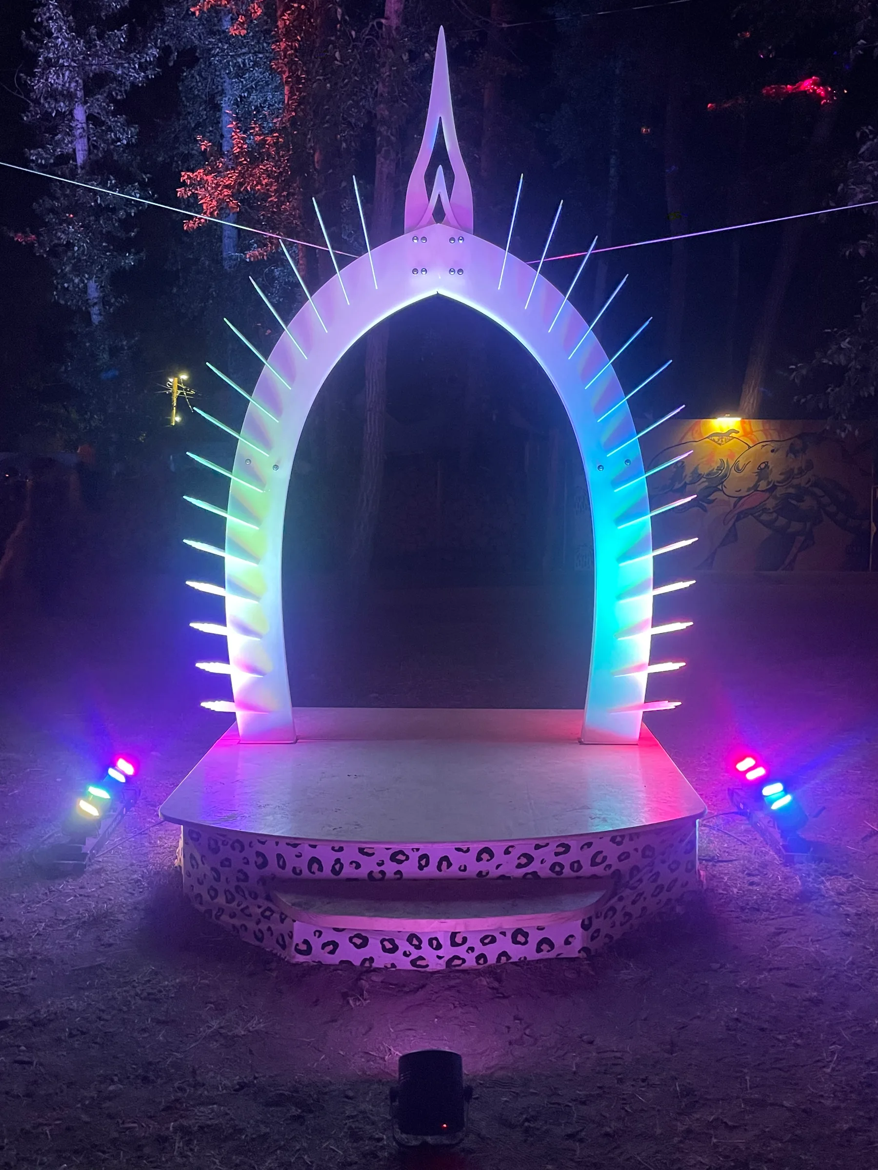 The Arch at night: its two sides rise and meet in a single point, glowing rainbow above the raised round platform in a dark forest