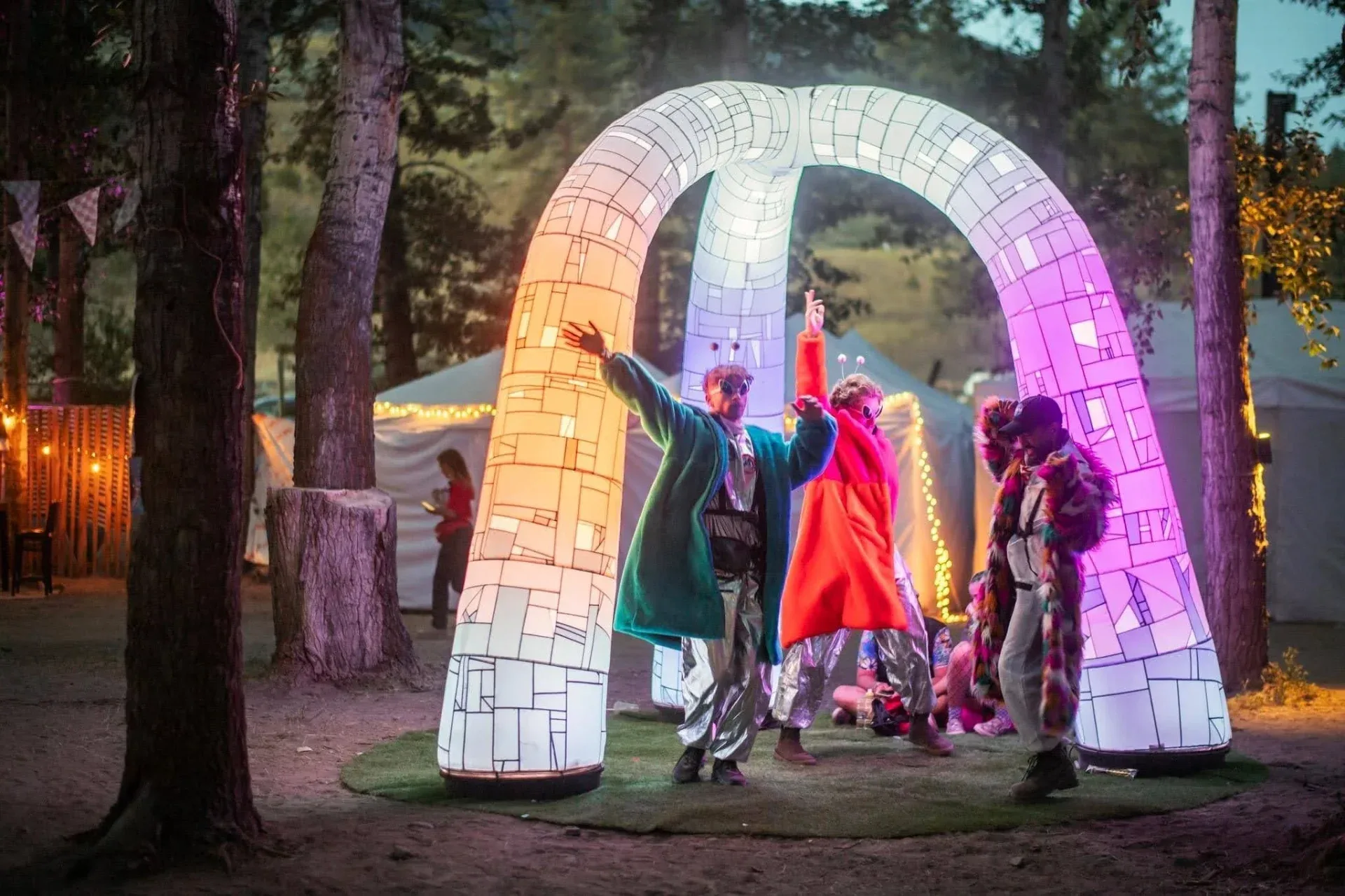 Puffy Portal glowing orange, white and purple at dusk among pine trees, with costumed dancers underneath