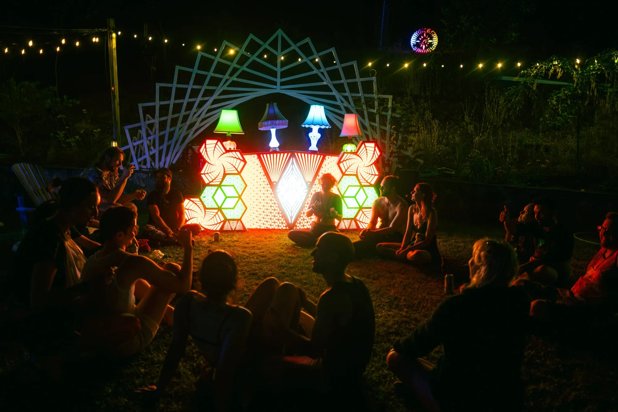 The Observer glowing orange and white at an outdoor night event, with people sitting on the grass in front