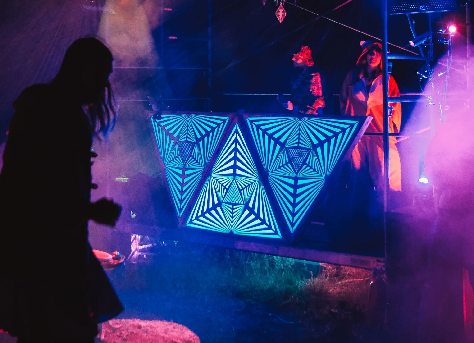 The Triangles glowing blue as the front of a DJ stage at a night festival, laser-cut starburst patterns lit through haze