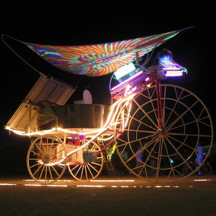 Daisy at night: a giant tricycle with glowing spoked wheels, lit frame and a painted sail over the passenger carriage