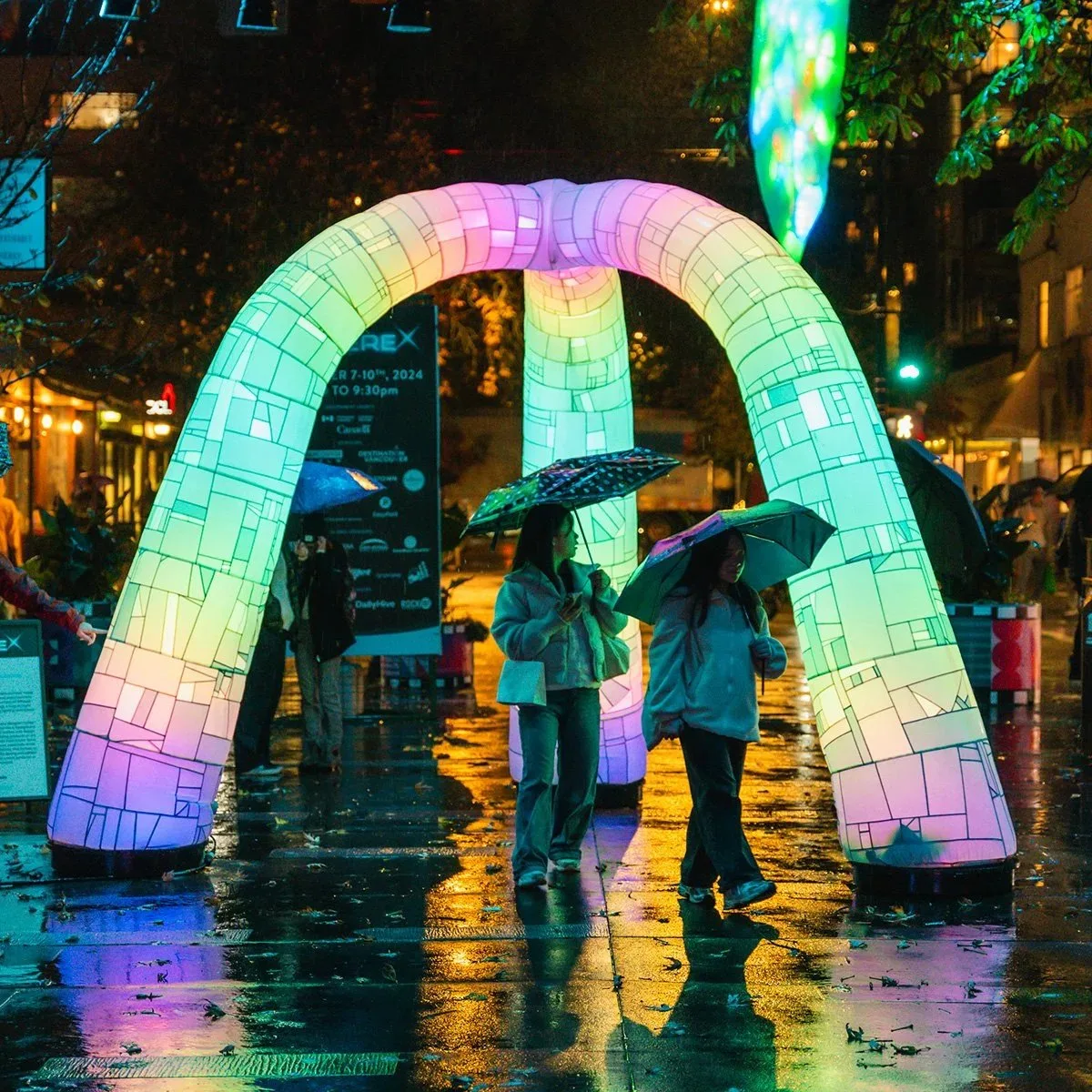 Puffy Portal glowing green, pink and violet on a rain-wet street at night, two people with umbrellas walking through it