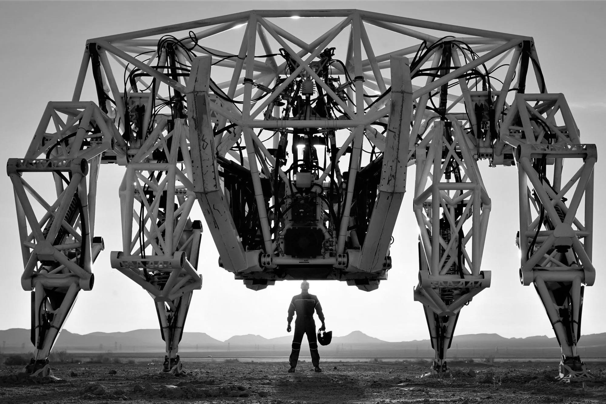 Prosthesis in black and white at dusk in the Mojave Desert, its pilot standing beneath the giant four-legged frame