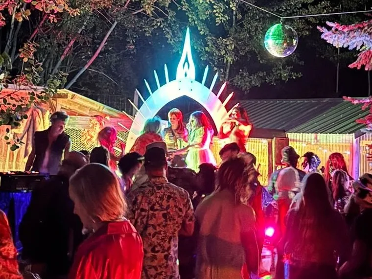 The Arch glowing at the end of a forest party at night, people dancing on its platform and a crowd gathered in front