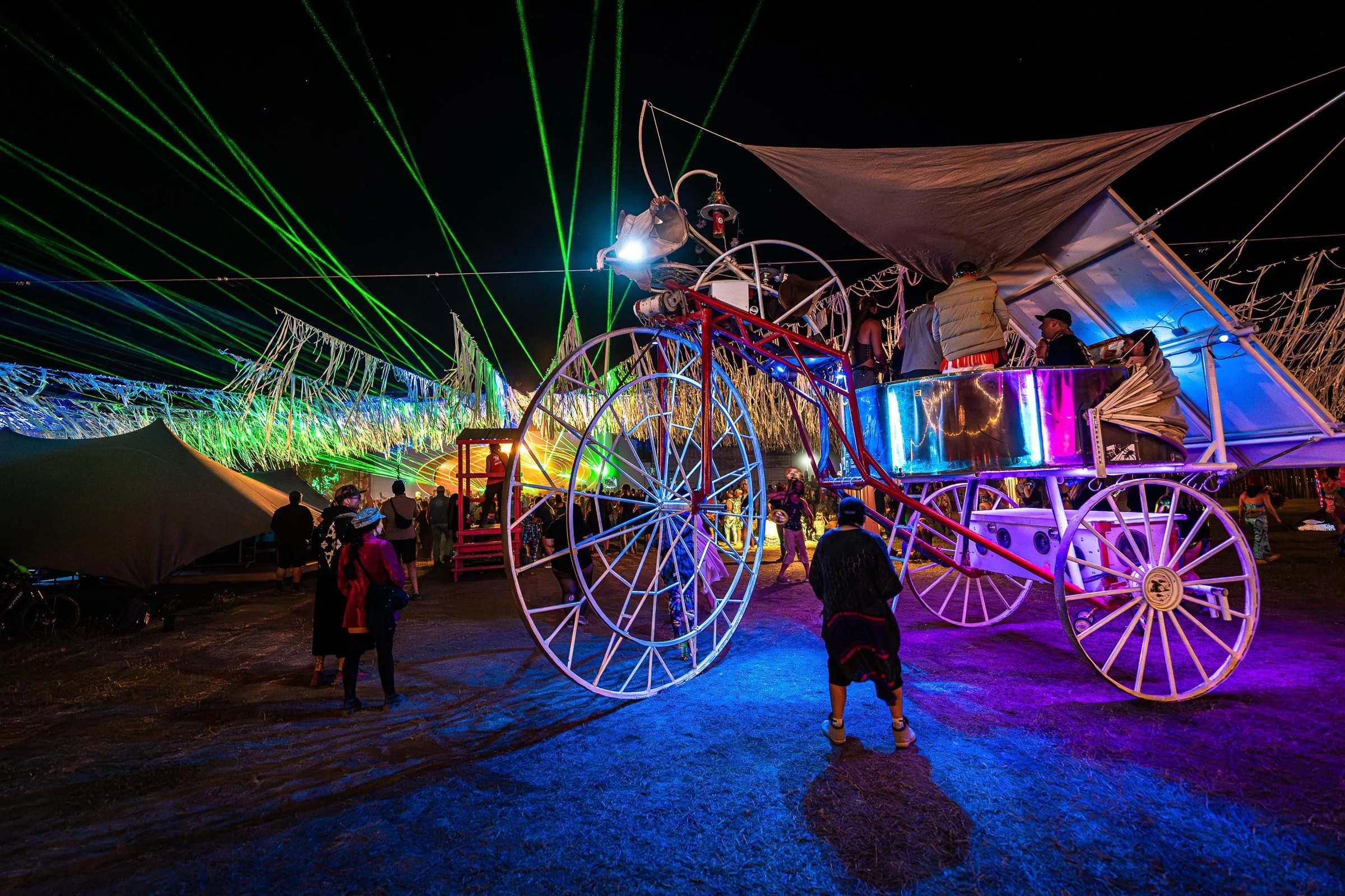 Daisy at night under green lasers, riders up in its lit carriage and people walking alongside the giant spoked wheels