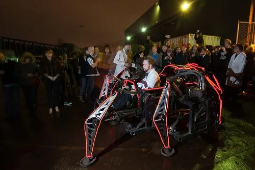 The Mondo Spider at night, its legs outlined in red light, with its rider aboard and a crowd gathered round