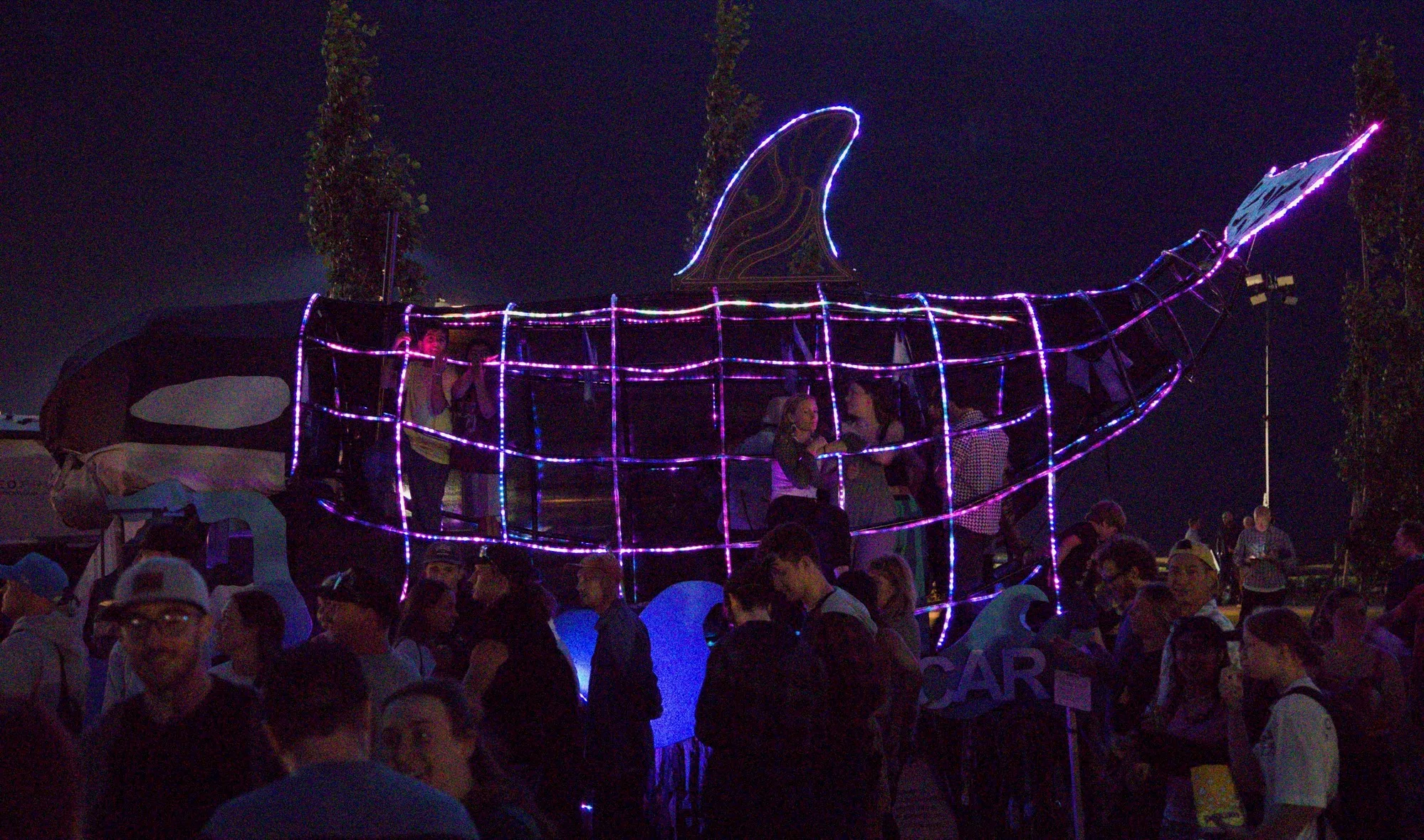 The Orcar at night lit in pink and blue LED rope, riders inside the ribs above a dense festival crowd